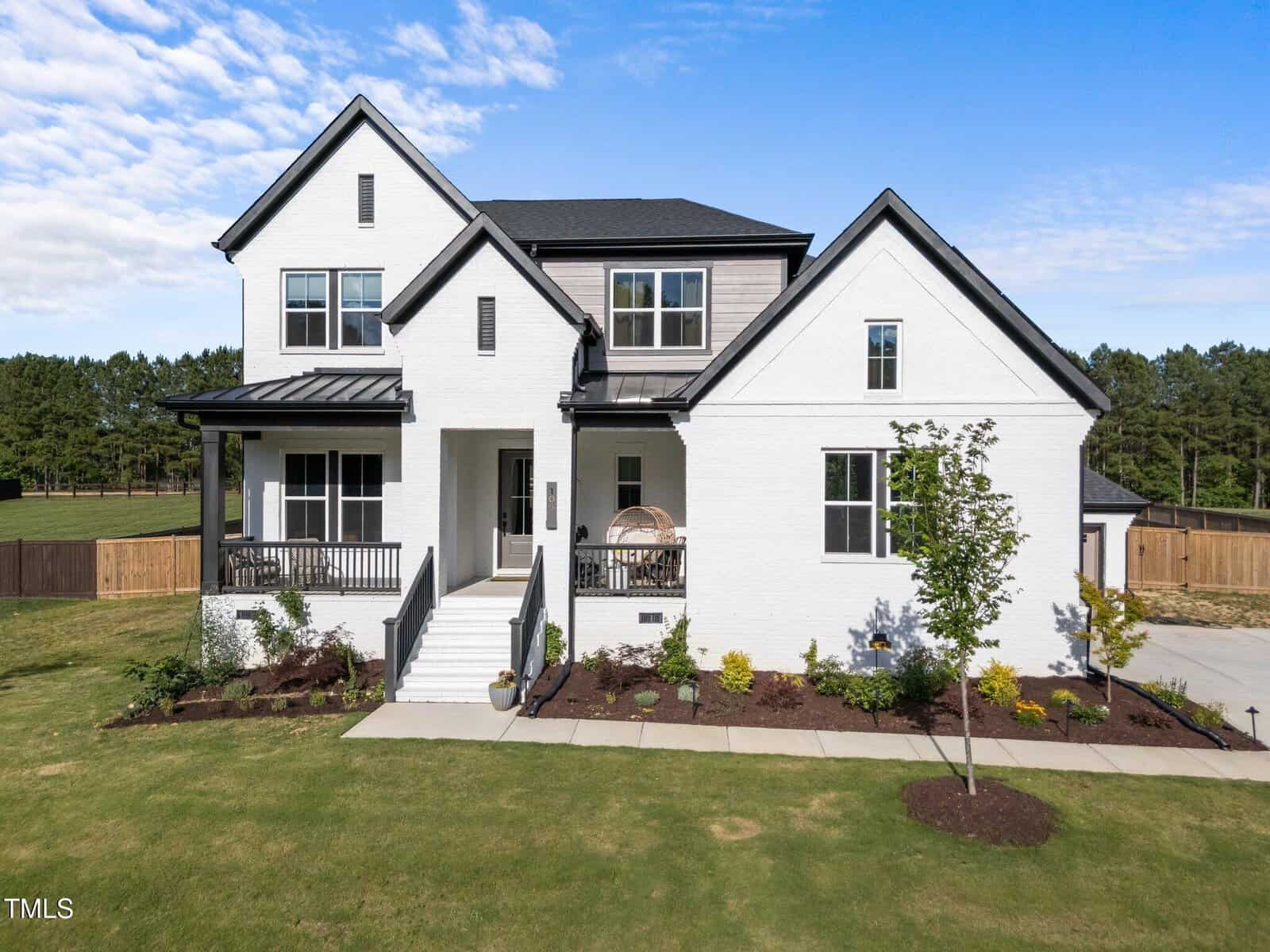 Modern two-story white brick house with a covered front porch, black trim, multiple gables, and a landscaped yard, set on a spacious lot with a wooden fence and surrounded by trees under a blue sky.