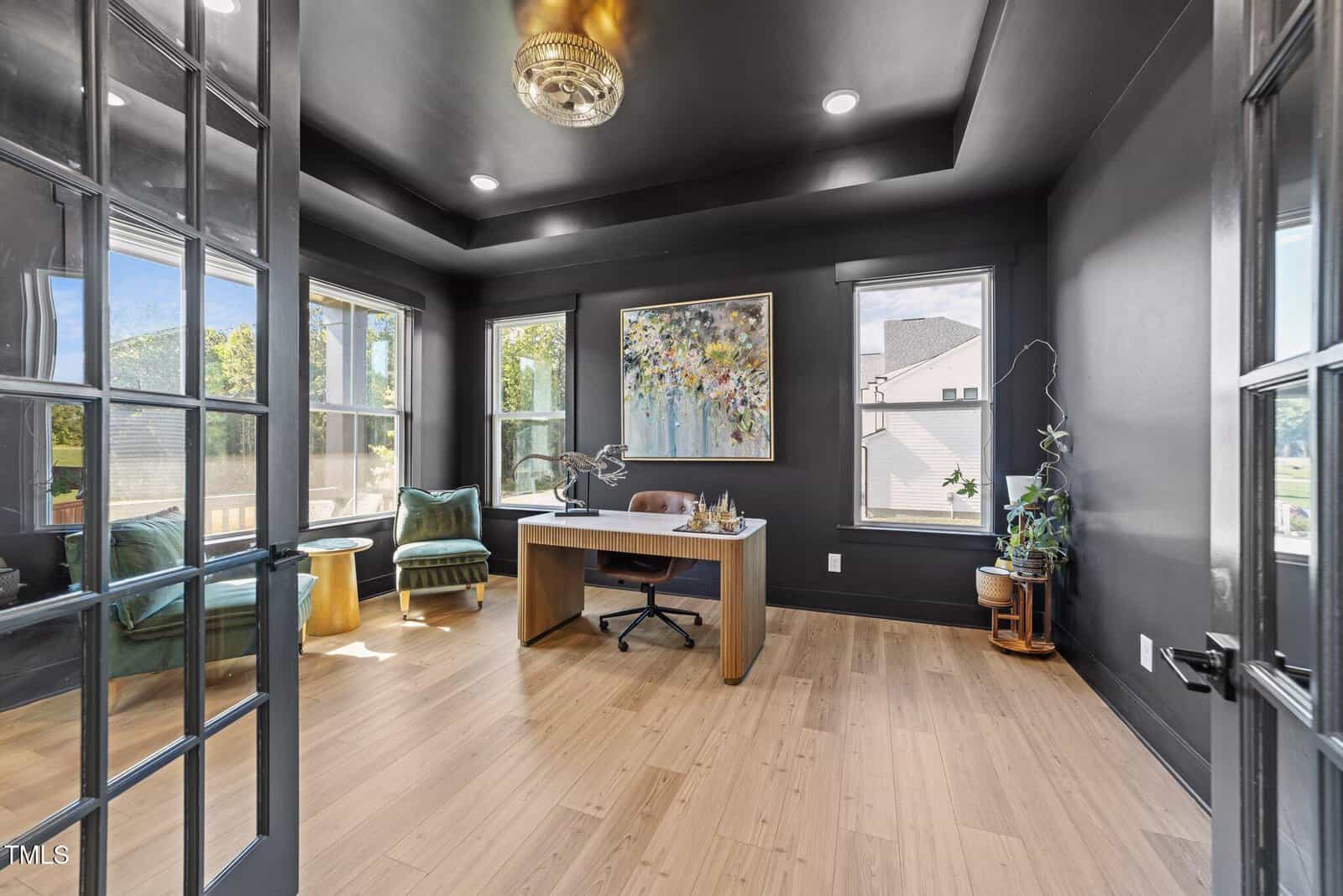 A modern home office with black walls, large windows, light wood floors, a central desk, green armchair, gold side table, potted plant, and colorful abstract artwork above the desk.