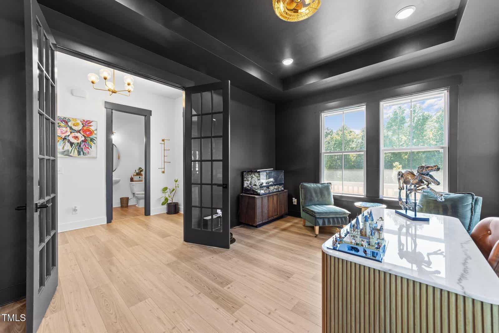 Modern home office with black walls and ceiling, light wood floors, double glass doors, aquarium, large windows, and a desk with a decorative bird sculpture. Bright bathroom with floral art is visible through open doors.