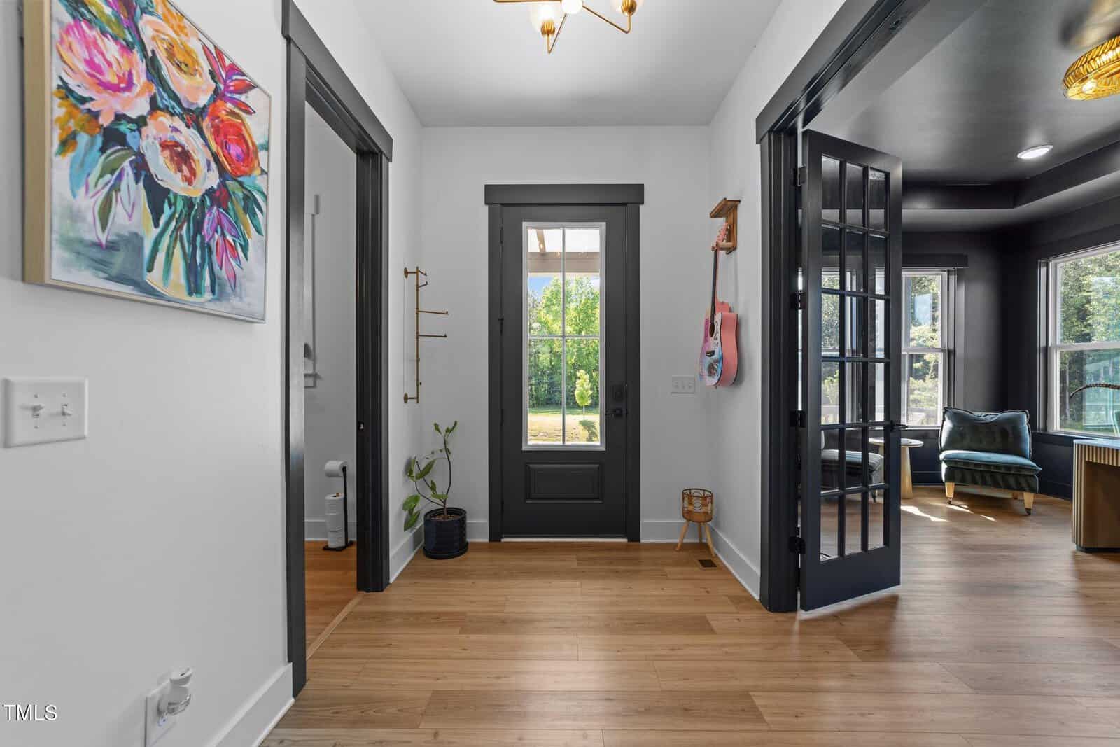 A bright entryway with light wood floors, a dark-framed glass door leading outside, and black-trimmed doors. A colorful floral painting hangs on the white wall, with a sunny room visible through French doors on the right.