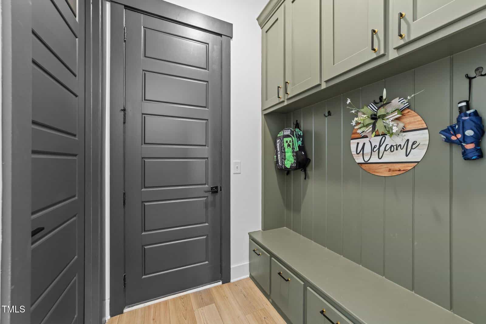 A mudroom with green built-in cabinets and bench, a gray paneled door, wood floor, a circular “Welcome” sign with flowers, a backpack, and blue rain boots hanging on hooks.