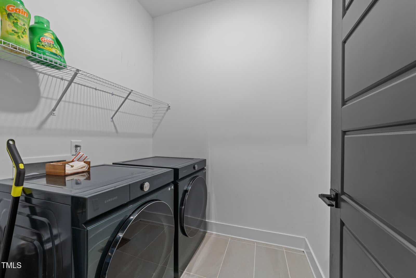 Modern laundry room with gray washer and dryer, a mop, and a wall shelf holding detergent bottles and a small wooden box, all against white walls and a light tiled floor.