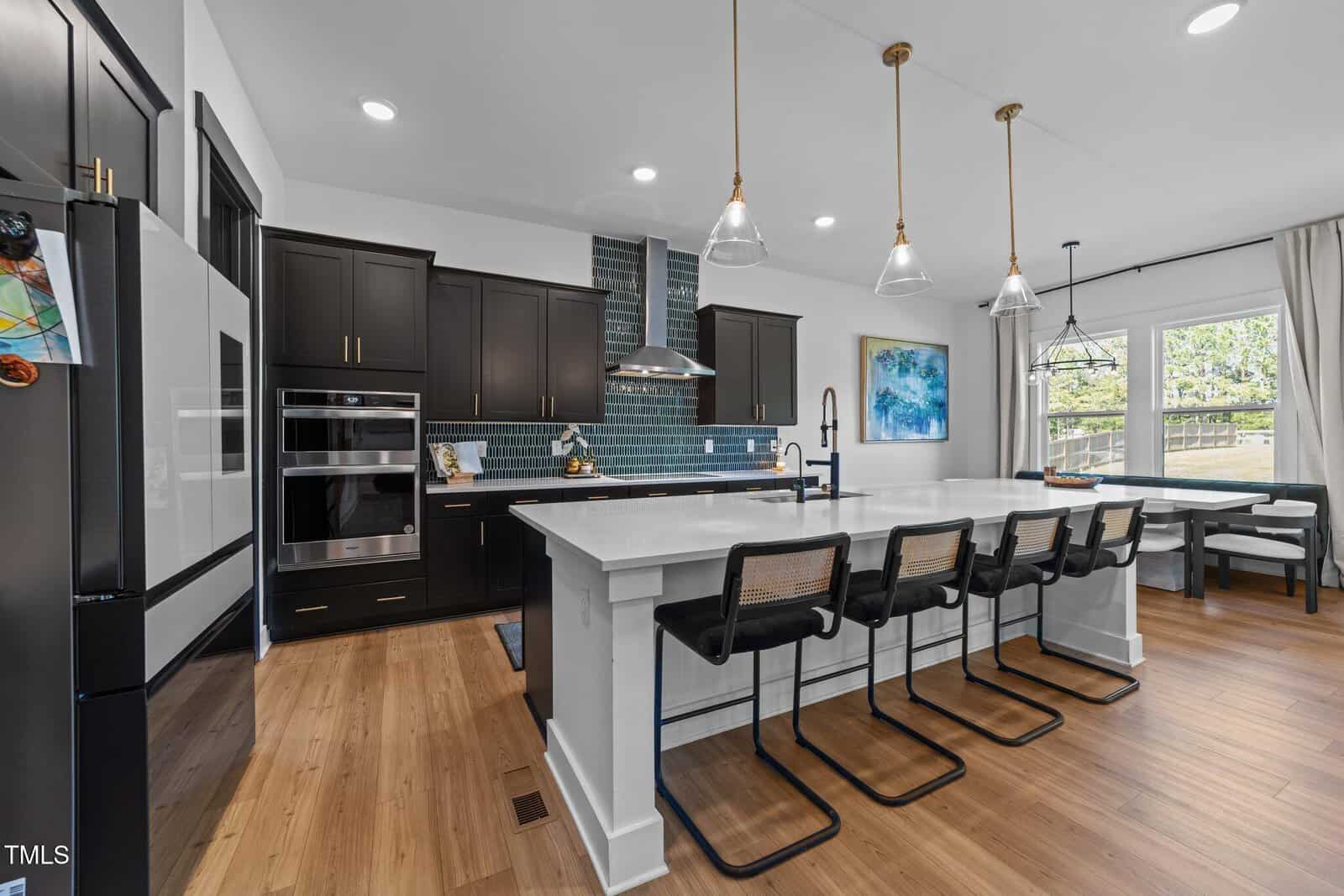 Modern kitchen with dark cabinets, a large white island with four barstools, stainless steel appliances, pendant lights, a blue tiled backsplash, and dining area with windows overlooking a backyard.