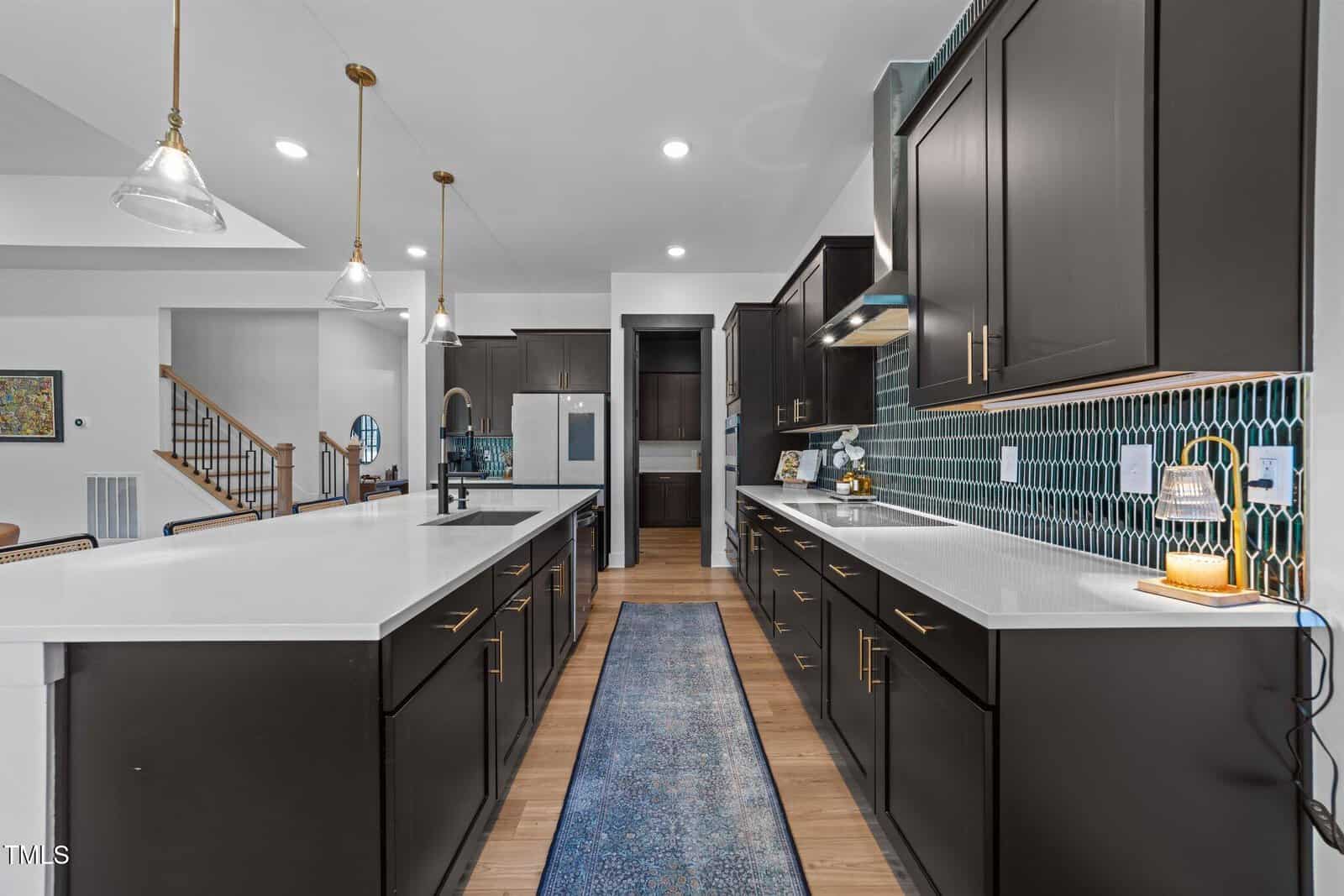 Modern kitchen with dark cabinets, white countertops, pendant lights, a blue rug, stainless appliances, and a tiled backsplash, viewed from the end of a central island.