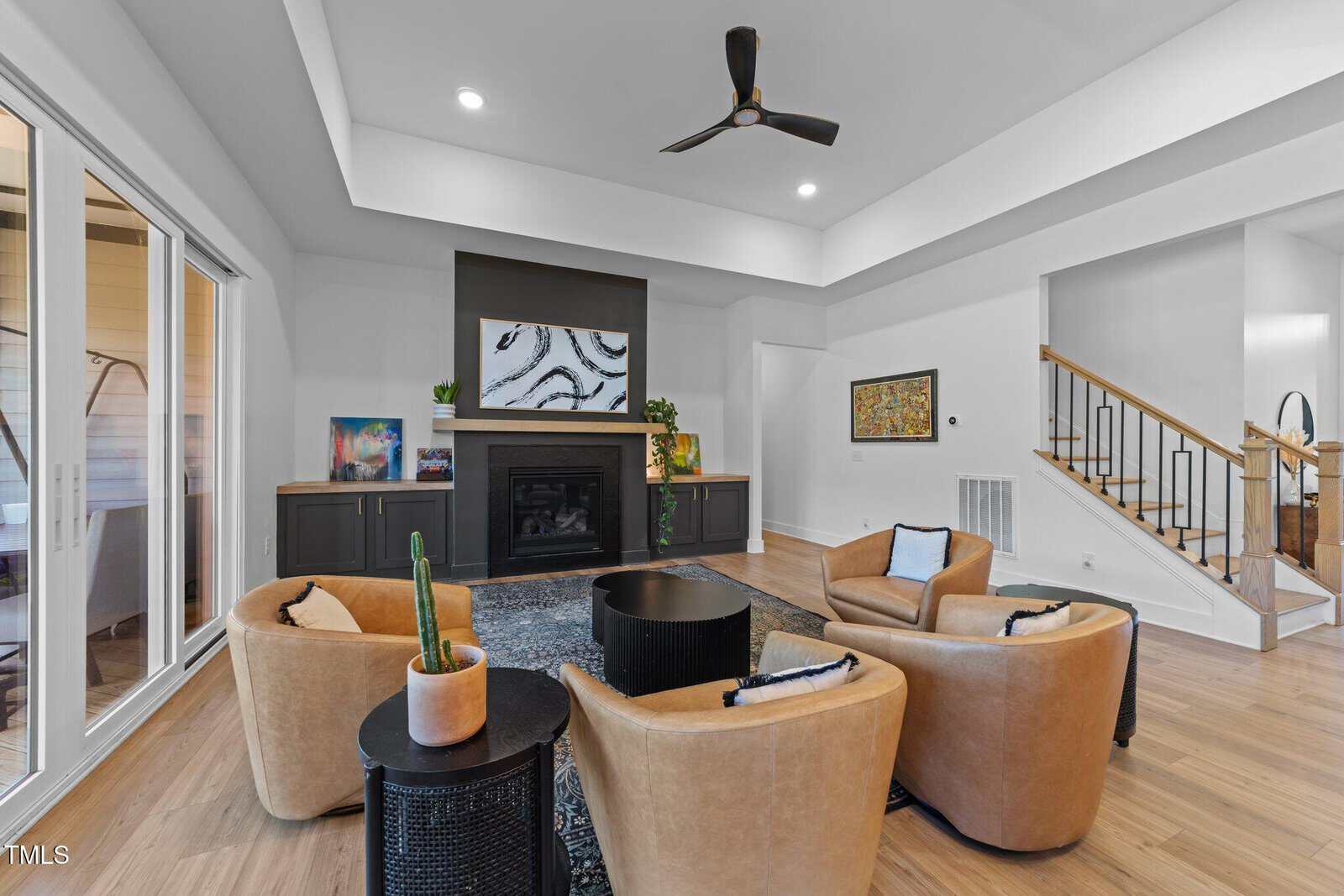 Modern living room with tan leather chairs, a round black table, a fireplace with art above it, and a ceiling fan. Light wood floors, white walls, and large windows create a bright, open space. Stairs are visible on the right.