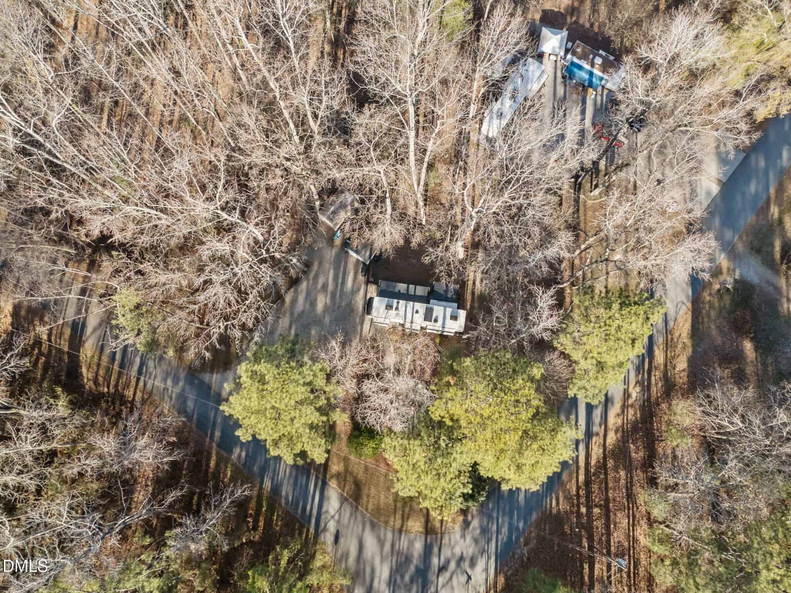 Aerial view of an RV parked in a clearing surrounded by leafless trees and some evergreen trees, with paved roads nearby and another small structure visible in the upper right corner. Shadows from trees fall across the scene.