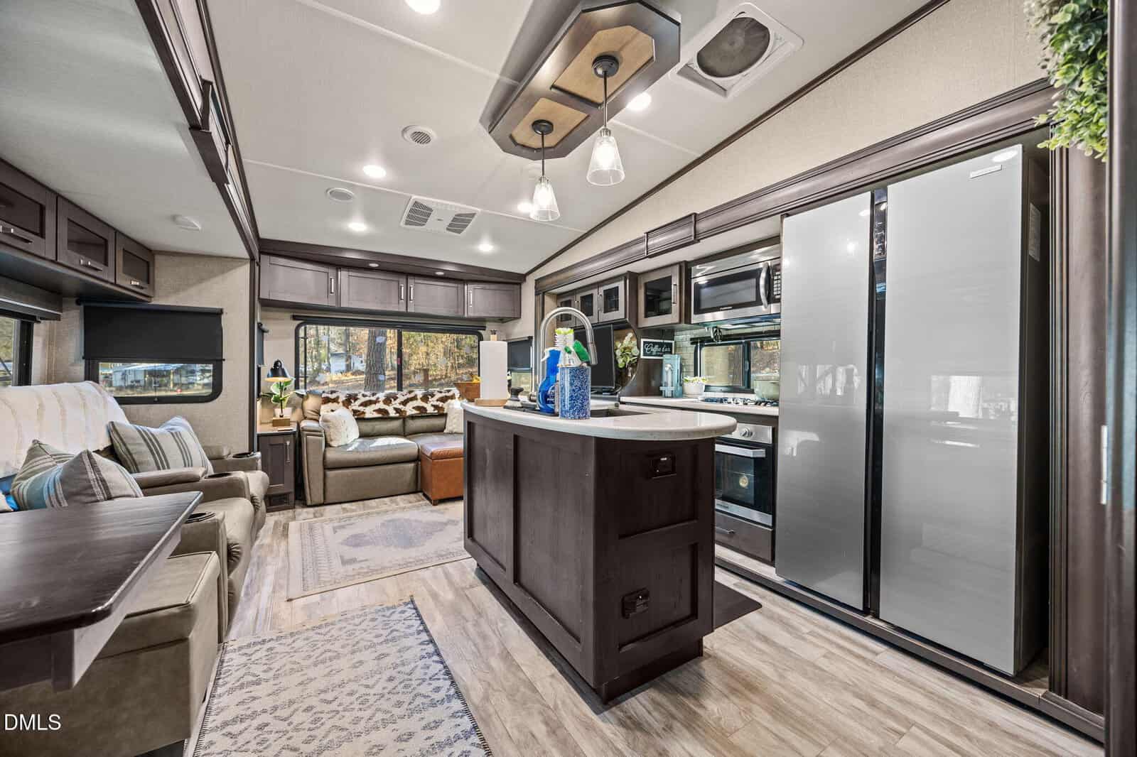 Modern RV interior at 115 Squaw Drive, Louisburg, featuring a kitchen island, double-door refrigerator, built-in microwave, sofa, recliner chairs, large windows with outdoor views, overhead cabinets, wood-style flooring, and sleek lighting fixtures.