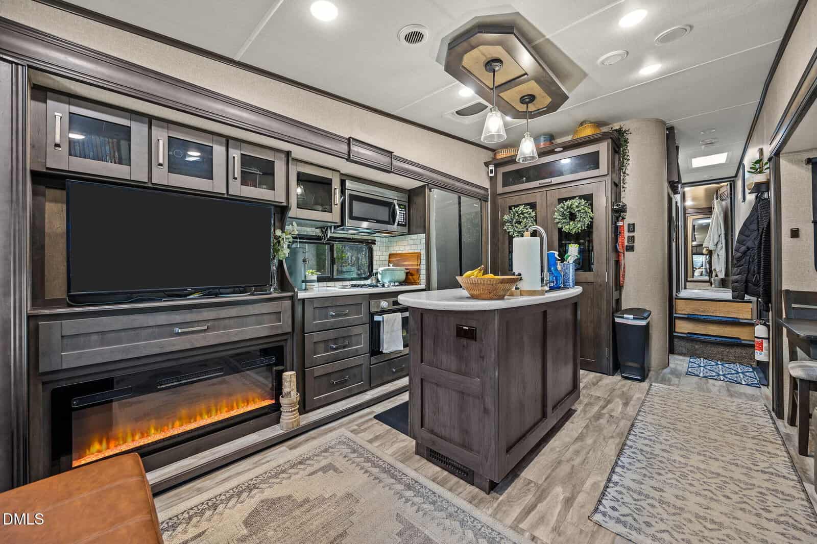 Modern RV interior featuring a kitchen island with a bowl of fruit, a large flat-screen TV above an electric fireplace, dark wood cabinetry, and steps leading to a raised area in the back.