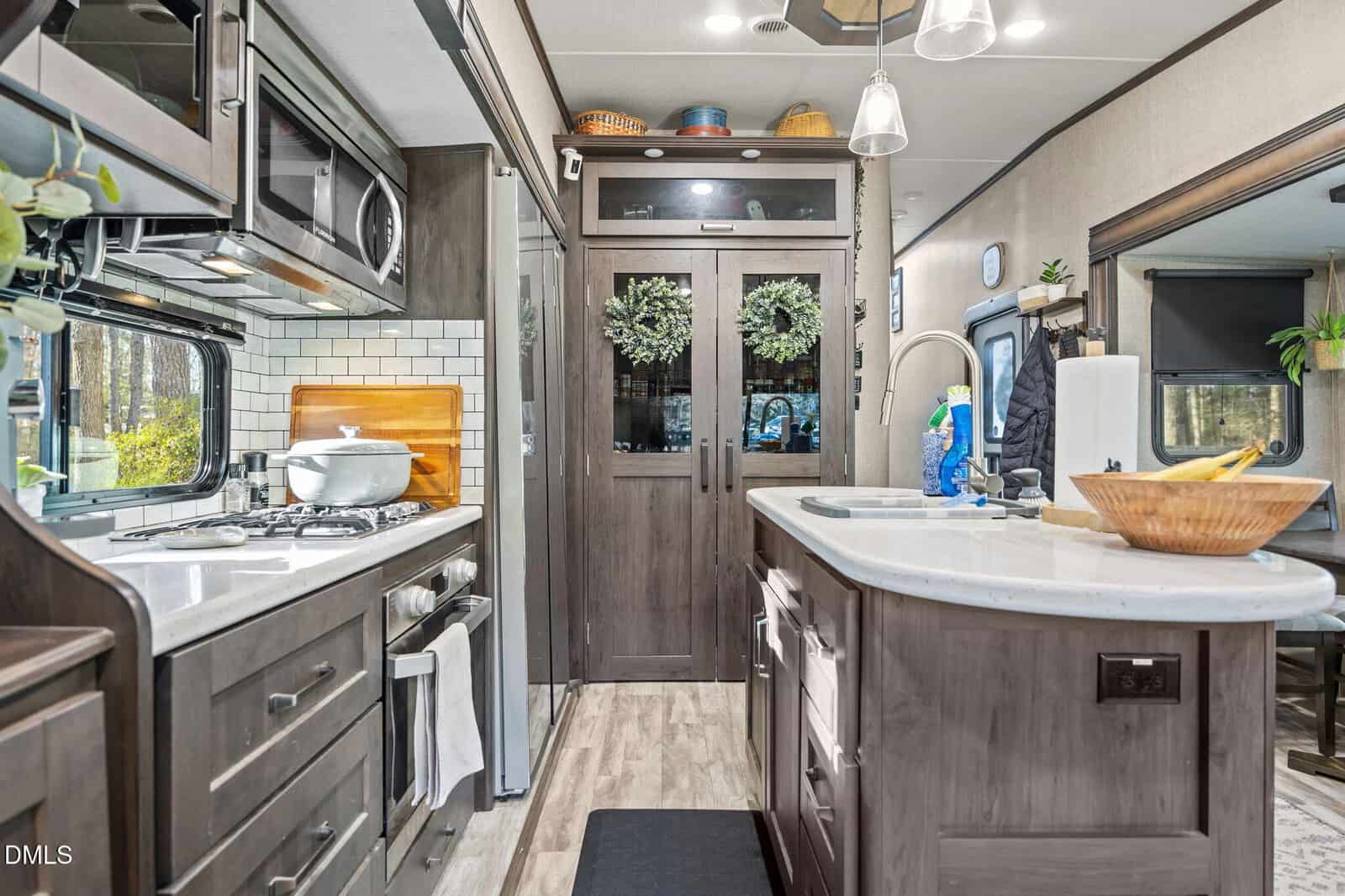 A modern RV kitchen with dark wood cabinets, a gas stove with a pot, white tile backsplash, microwave, and a center island with a sink. Double glass doors with wreaths are at the end of the kitchen.