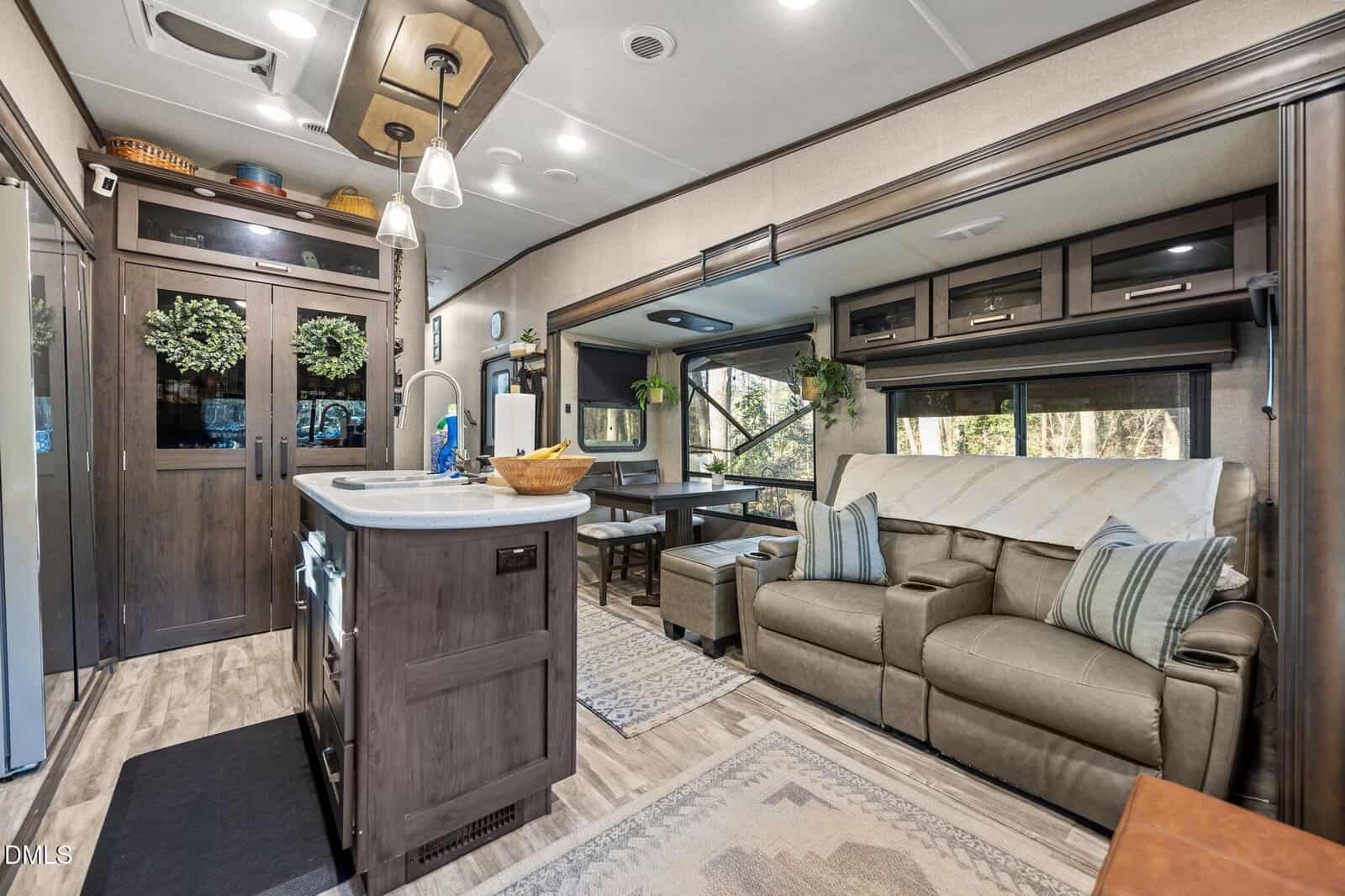 Modern RV interior with a kitchen island, dining nook, and beige sofa. Neutral tones, wood cabinets, area rug, and large windows with outdoor views create a cozy, inviting living space.