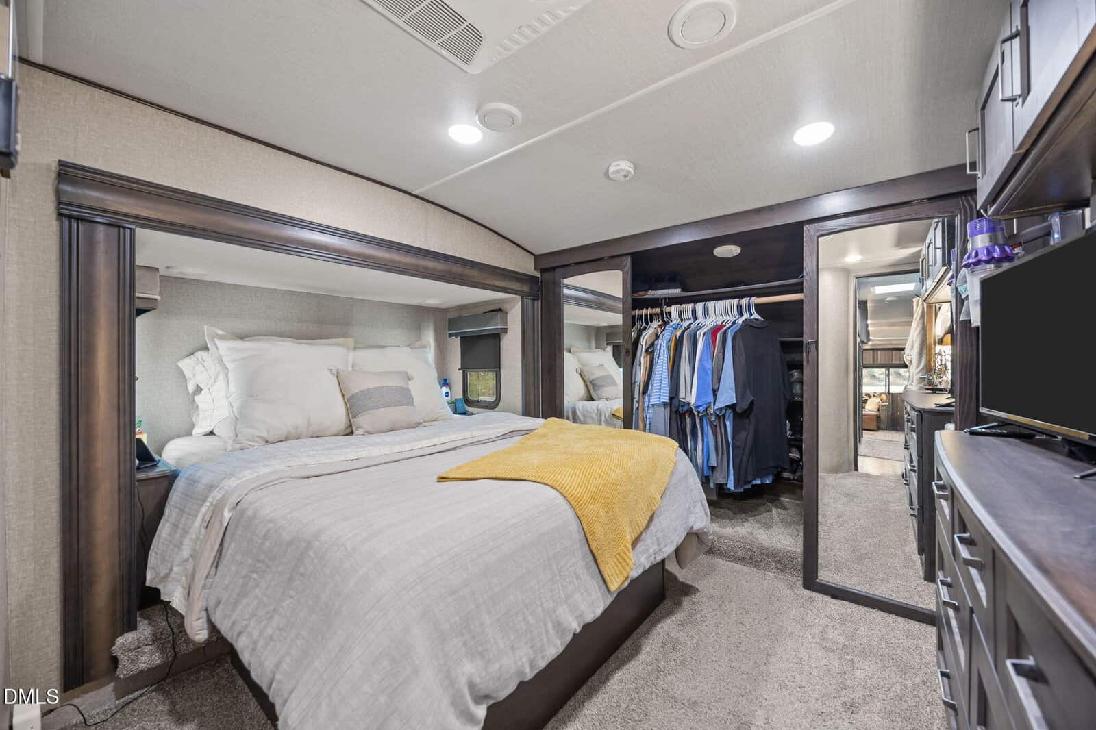 A neatly made bed with neutral bedding and a yellow throw sits next to a mirrored closet with hanging clothes in a modern, carpeted bedroom. A dresser with a TV is visible, and natural light comes from a window.
