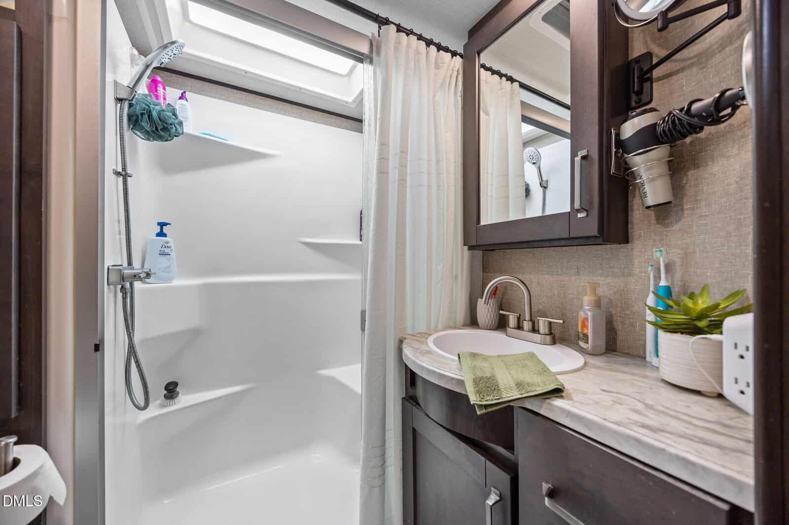 A compact bathroom features a shower with shelves, a white sink with a green towel, a marble-patterned countertop, dark wood cabinets, a hair dryer, toiletries, and a small potted plant. A skylight is above the shower.