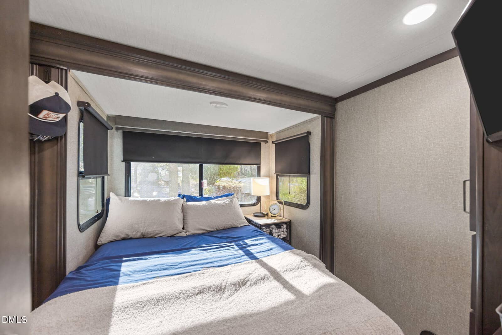 A compact bedroom with a double bed, blue and white bedding, two windows with blackout shades, a small nightstand with a lamp, and a baseball cap hanging on the wall.