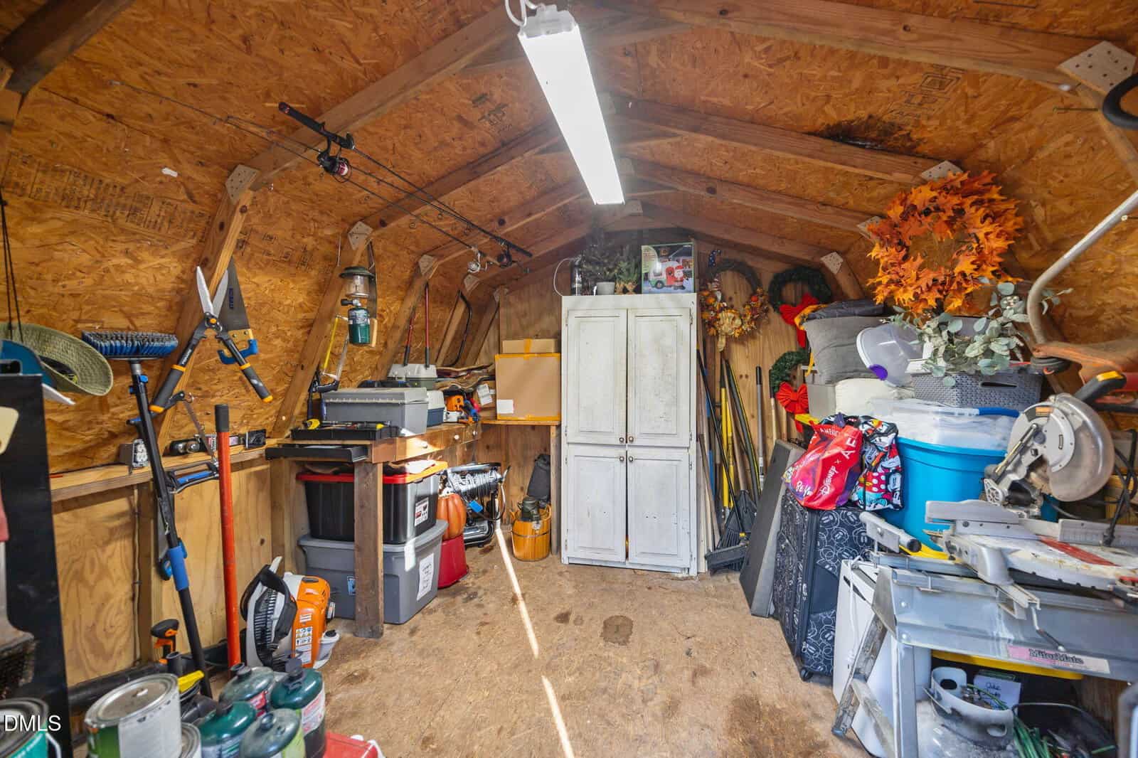 Interior of a cluttered wooden shed with tools, storage bins, a white cabinet, artificial plants, a saw, and various gardening items organized along the walls and floor under a fluorescent light.
