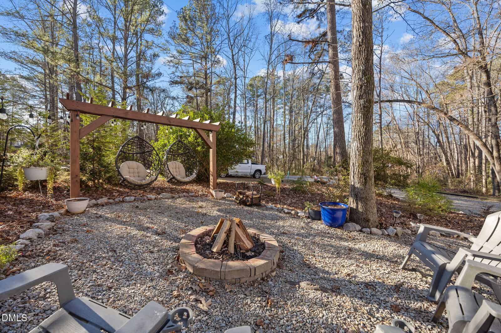 A gravel patio with a fire pit, surrounded by chairs and two hanging basket seats under a wooden pergola, set in a wooded area with tall trees and a white pickup truck in the background.