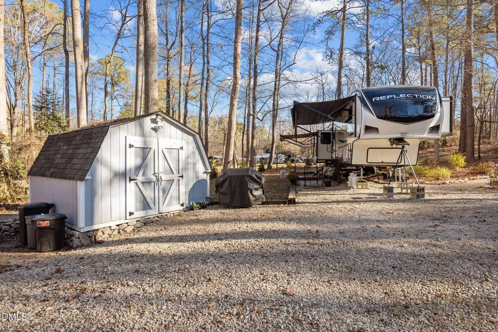 A white fifth-wheel RV trailer on cinder blocks is parked beside a small gray shed with double doors, surrounded by tall trees and gravel, under a partly cloudy sky. A covered grill and trash bins are nearby.