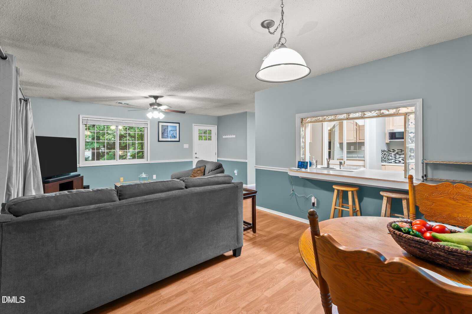 A living room with gray sofas faces a TV in the corner, adjacent to a dining area with a wooden table and chairs. A breakfast bar with stools separates the kitchen, and large windows let in natural light.