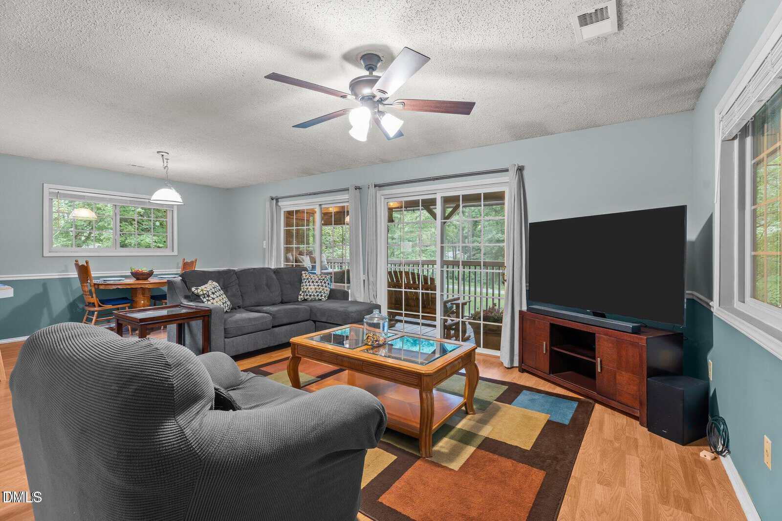 A living room with a gray sofa, matching armchair, glass coffee table, and flat-screen TV on a wooden stand. Sliding glass doors with curtains open to a covered porch. A small dining area is visible in the corner.