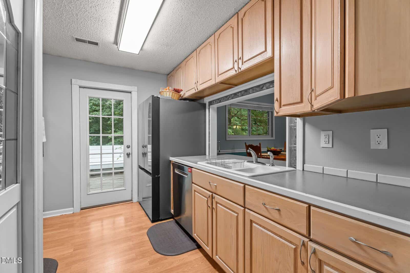 A kitchen with light wood cabinets, white tile countertops, stainless steel appliances, and wood flooring. There is a double sink, a window opening to the dining area, and a glass door leading outside.