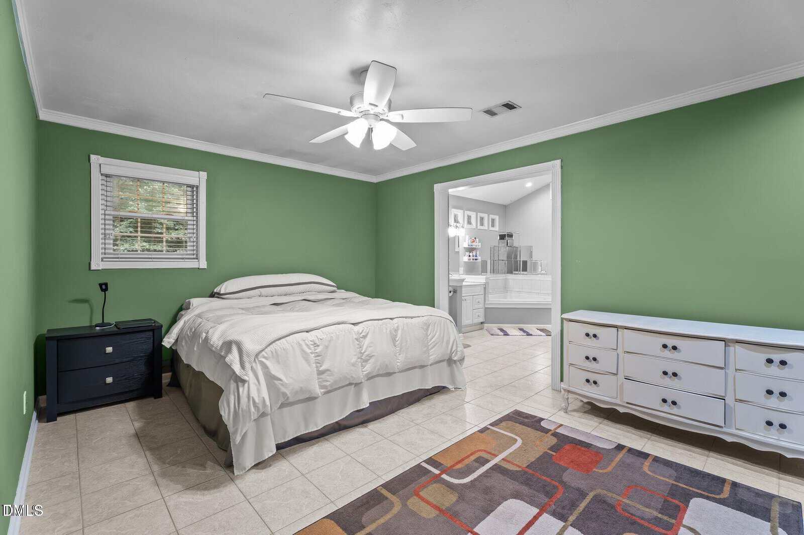 Bedroom with green walls, white ceiling fan, bed with white bedding, two black nightstands, white dresser, colorful rug, and an open doorway leading to a bathroom with a tub and white cabinets.