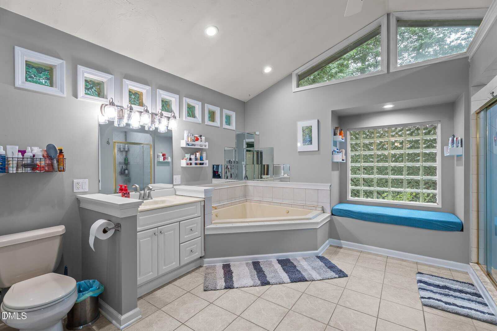 Spacious bathroom with a large bathtub, glass shower, single sink vanity, toilet, and window seat under tall windows. Gray walls and light tile floor with rugs give a clean, modern feel.