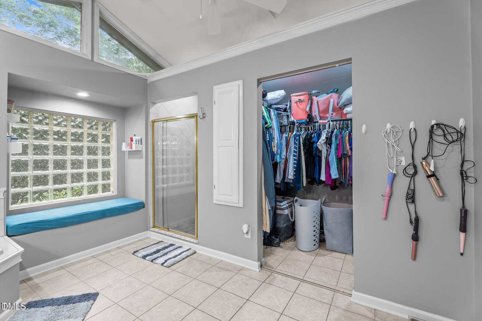 A bright bathroom with a glass shower, window seat, tile floors, open closet with clothes, and hair tools hanging on the wall. A laundry basket sits in the closet; large windows let in natural light.