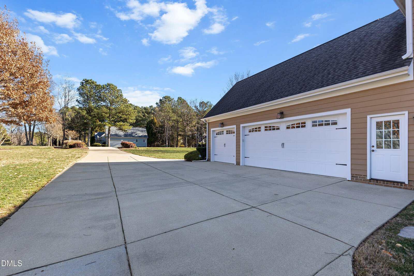 A wide concrete driveway leads to a three-car garage attached to a tan house with white trim and a side door, surrounded by grass, trees, and a clear blue sky.