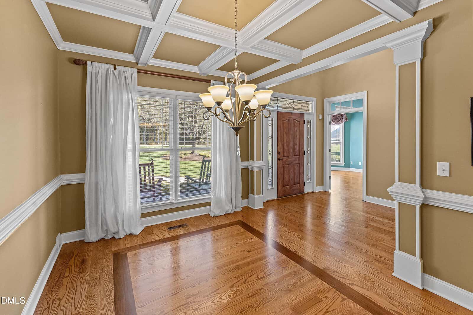 A dining room with hardwood floors, beige walls, white ceiling beams, a central chandelier, large windows with curtains, and a wooden front door; an adjacent room with blue walls is visible.