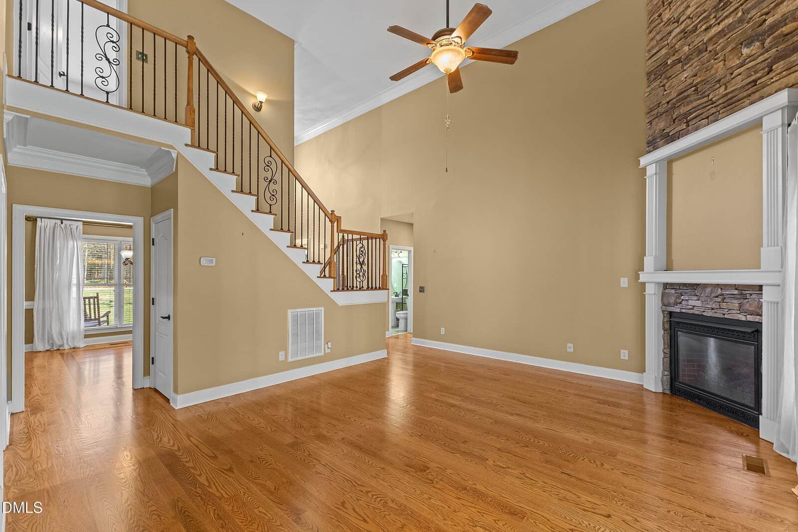 Spacious living room with wood floors, high ceiling, ceiling fan, stone fireplace, and staircase with metal railings. Doorways lead to other rooms with large windows and natural light.