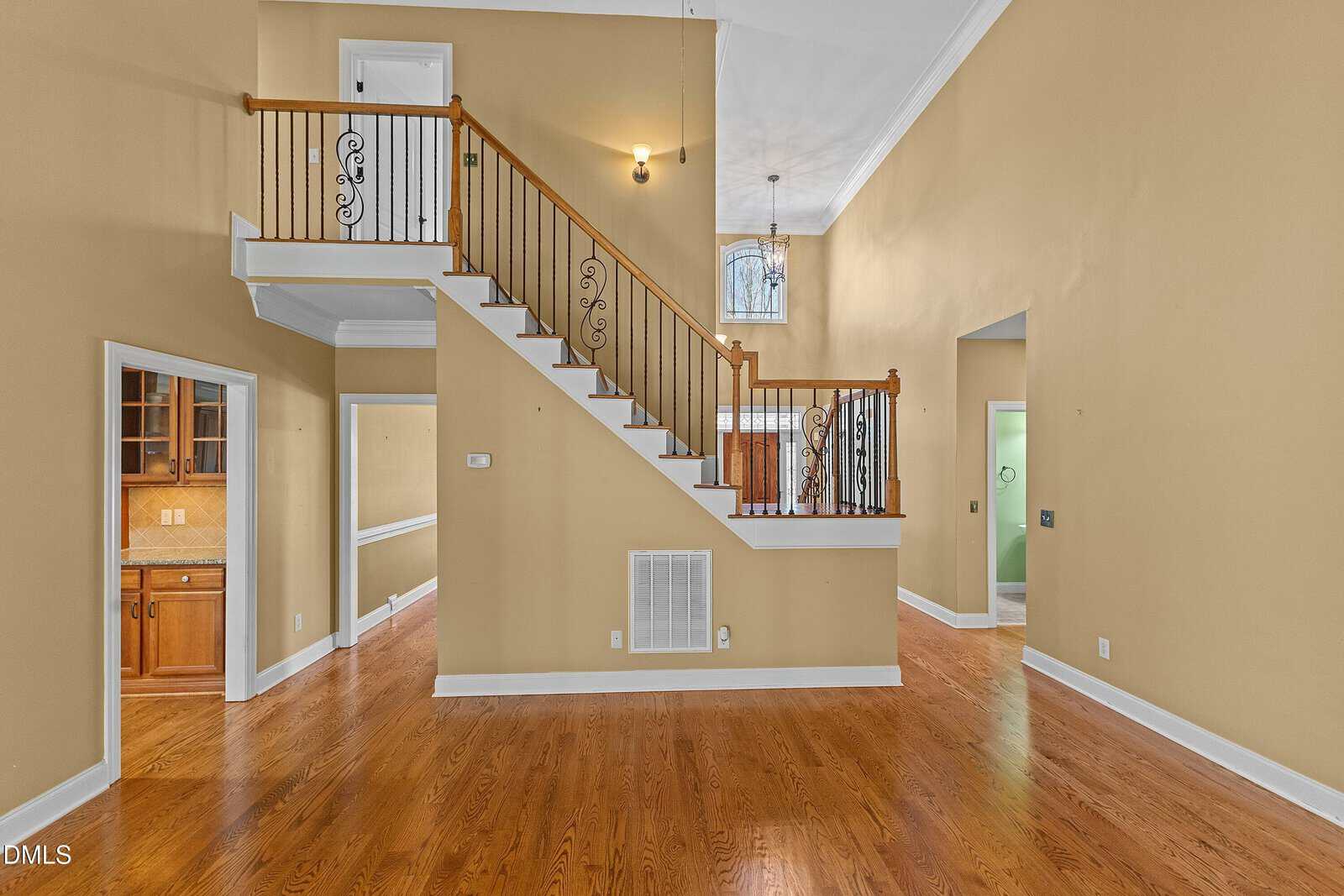 Spacious room with wood floors, yellow walls, a staircase with a metal railing, and a high ceiling. Light fixtures and doors to other rooms are visible. Natural light enters from a window above the front door.