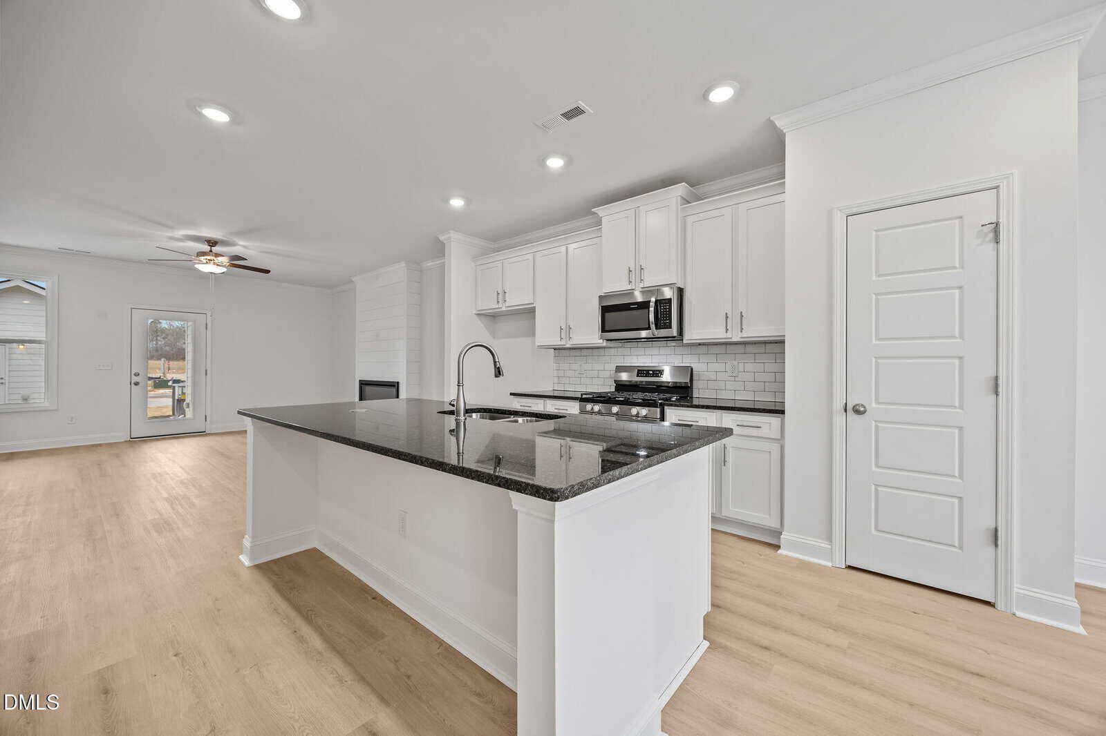 Modern kitchen with white cabinets, stainless steel appliances, a large black granite island with sink, subway tile backsplash, and light wood flooring, opening into a spacious living area with ceiling fan and windows.