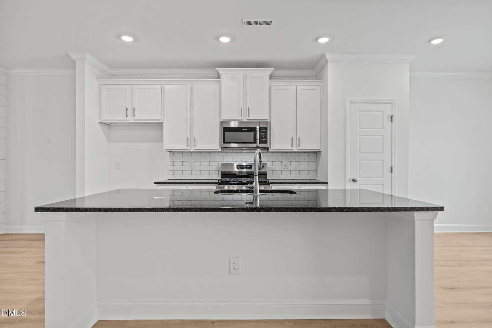 Modern kitchen with white cabinets, stainless steel appliances, a subway tile backsplash, and a large black granite island countertop with a built-in sink and faucet. Bright, minimalistic design with light wood flooring.