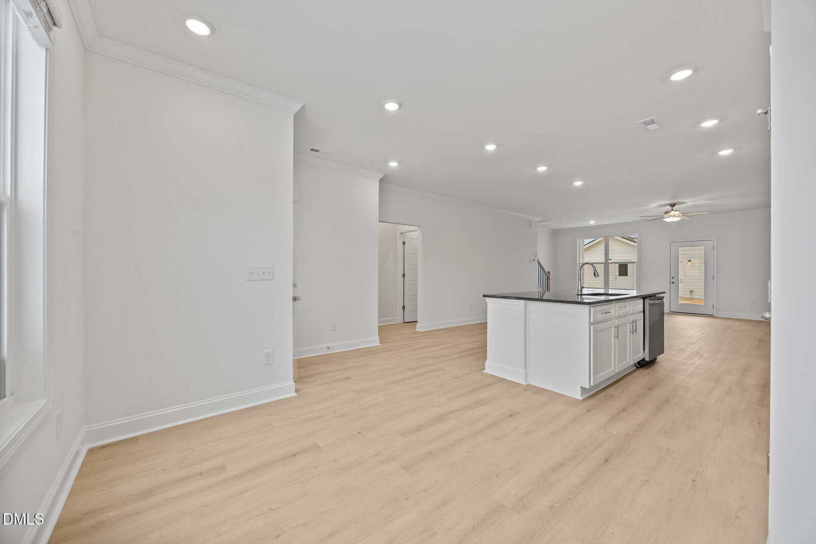 Spacious, modern kitchen and living area with light wood flooring, white walls, recessed lighting, center island with sink, and large windows providing natural light. Door leads to an outdoor area.