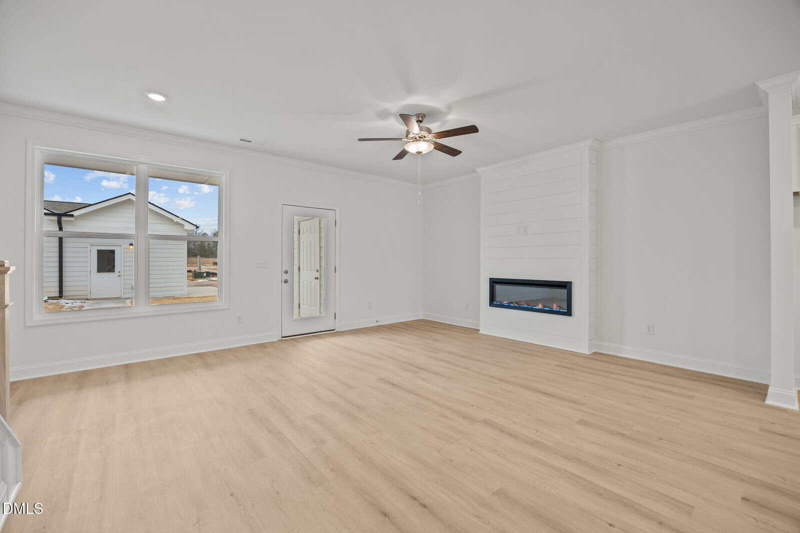 Spacious, empty living room with light wood floors, white walls, a ceiling fan, large window, glass door, built-in modern fireplace, and view of neighboring house and blue sky outside.