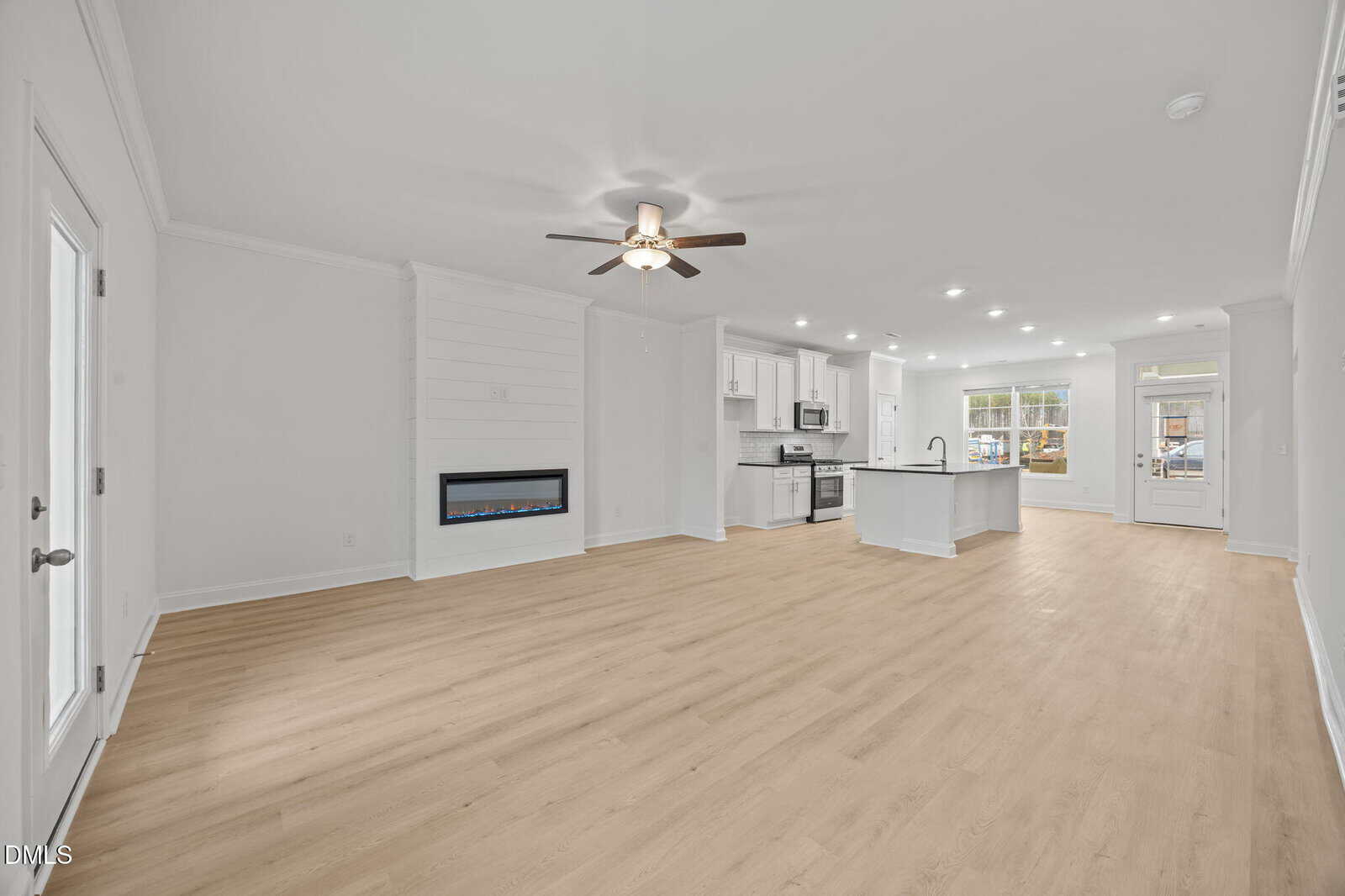 Open-concept living space with light wood floors, white walls, a ceiling fan, built-in electric fireplace, and a modern kitchen with an island, stainless steel appliances, and recessed lighting. Large windows let in natural light.