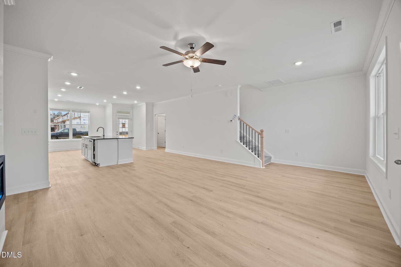 Open-concept living area with light wood floors, white walls, ceiling fan, kitchen with island and stainless appliances, large windows, recessed lighting, and a staircase leading upstairs.