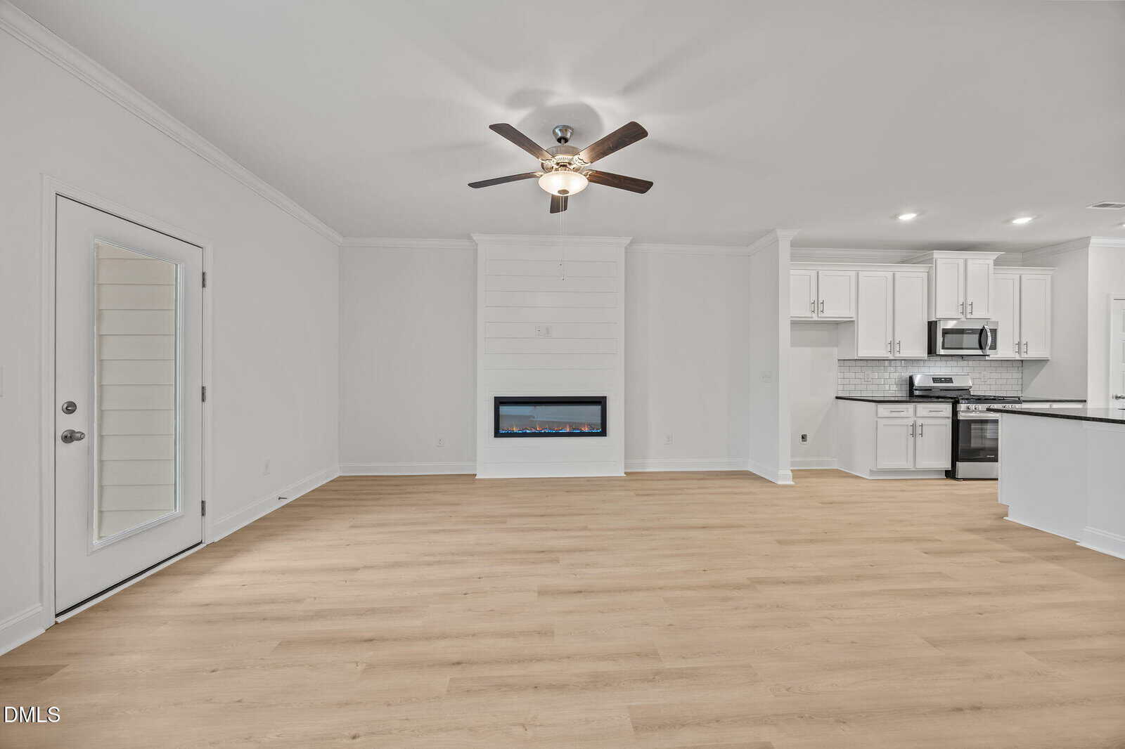 Modern, bright living area with light wood flooring, white walls, a ceiling fan, electric fireplace, glass patio door, and an open kitchen with white cabinets, stainless steel appliances, and black countertops.