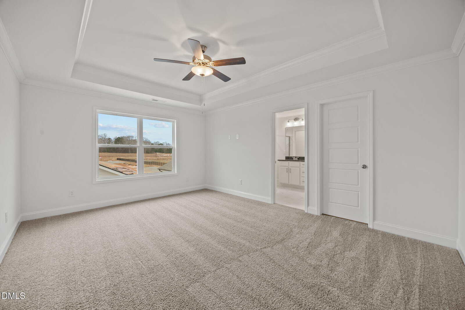 A spacious, empty bedroom with beige carpet, a tray ceiling with a ceiling fan, a large window, and a door leading to an en-suite bathroom with a double-sink vanity.