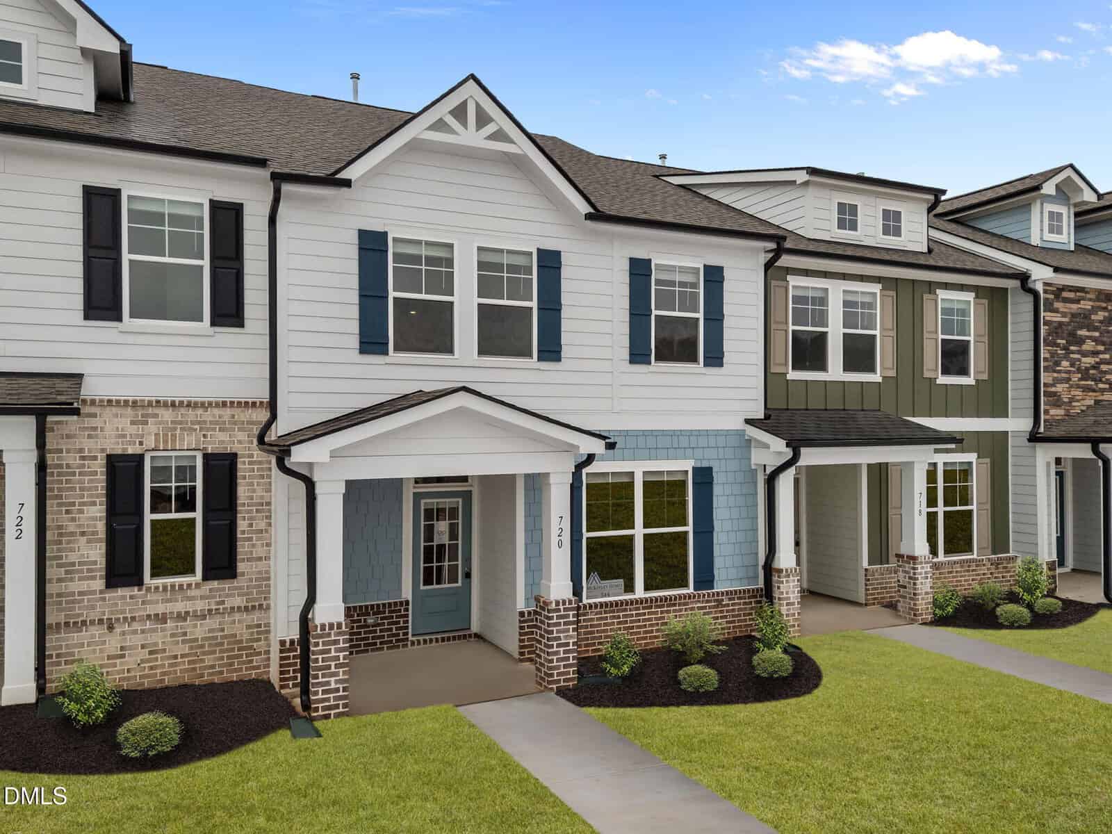 A row of modern townhouses with brick and siding exteriors, each featuring a small front porch, manicured lawns, and neatly trimmed shrubs under a bright blue sky.