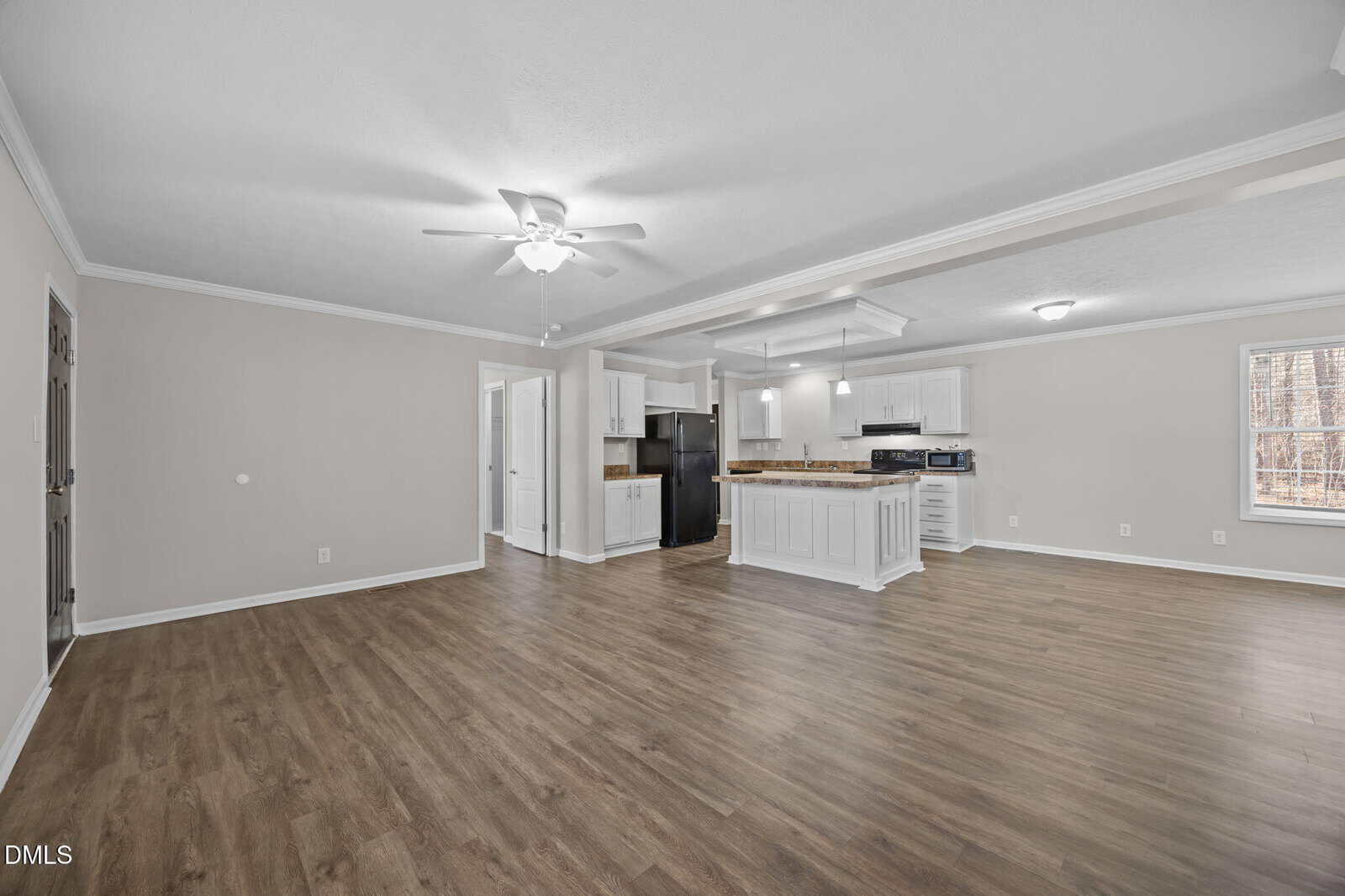 Spacious open-concept living area with light gray walls, wood-style flooring, ceiling fan, and a modern kitchen featuring white cabinets, an island, black appliances, and a window letting in natural light.