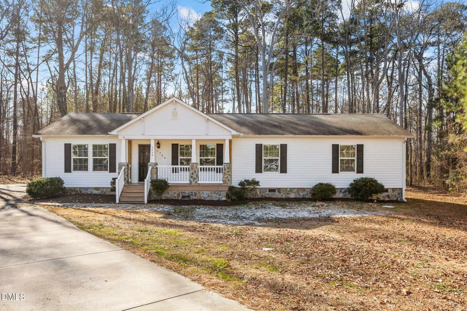 A single-story white house with a covered front porch, black shutters, and a gabled roof, set against a backdrop of tall trees with a driveway and patchy grass in the front yard.