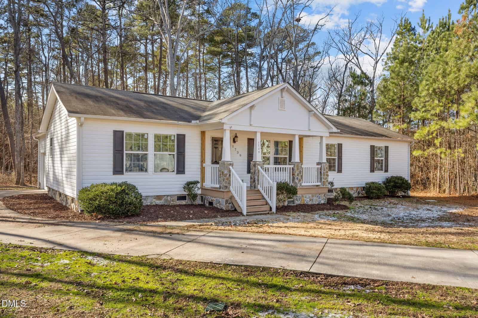 Single-story white house with gray shutters and a covered front porch, surrounded by trees and shrubbery, on a clear day with a blue sky and scattered clouds.