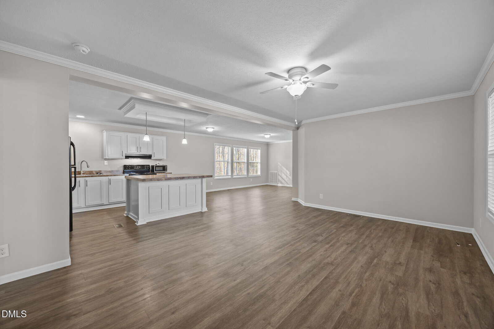 Spacious open-plan living area with wood flooring, white walls, and ceiling fans. The kitchen features white cabinets, an island, and stainless steel appliances. Large windows allow natural light into the space.