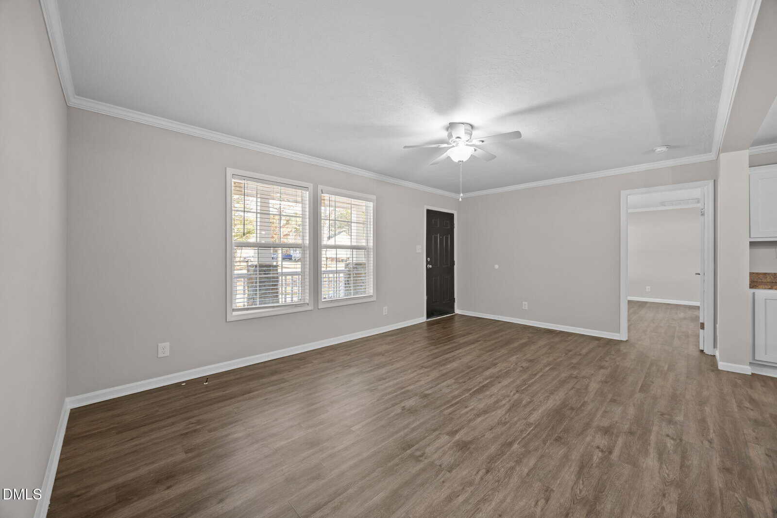 A bright, empty living room with light gray walls, wood-look flooring, large windows, a ceiling fan, and an open doorway leading to another room.