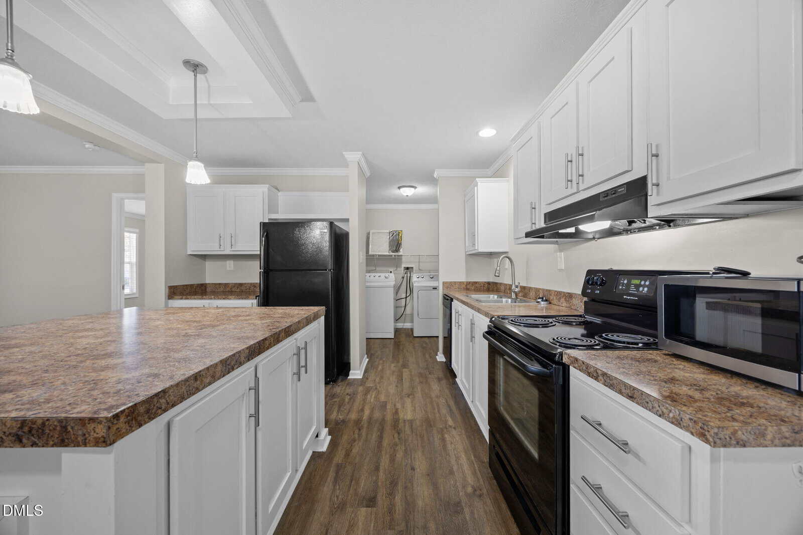 Modern kitchen with white cabinets, dark appliances, wood-style flooring, and brown countertops. In the background, a laundry area with a washer and dryer is visible through an open doorway.