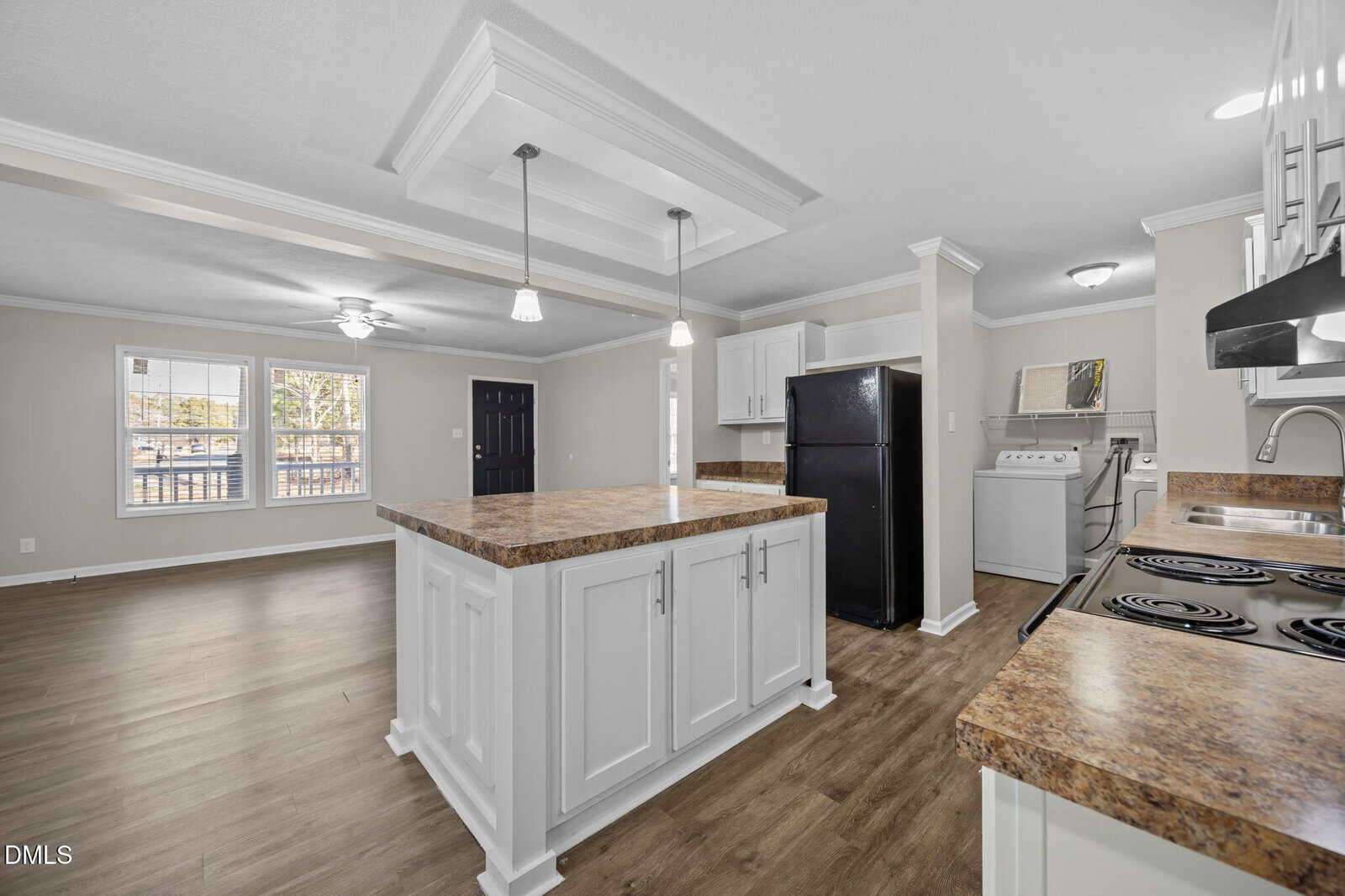 Modern open kitchen with white cabinets, brown countertops, an island, black appliances, and an adjacent laundry area with washer and dryer. Wood flooring extends into a bright living space with large windows.