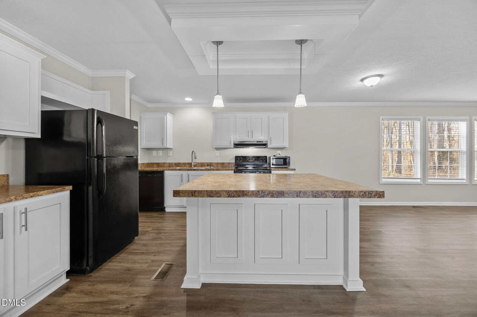 Modern kitchen with white cabinets, a large center island, black appliances including a refrigerator and stove, brown countertops, pendant lights, and large windows letting in natural light.