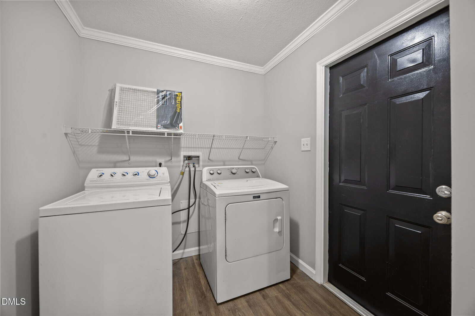 Small laundry room with a white washing machine and dryer, a black door, and a wire shelf holding a box and white basket. The walls are gray, and the floor is wood-patterned.
