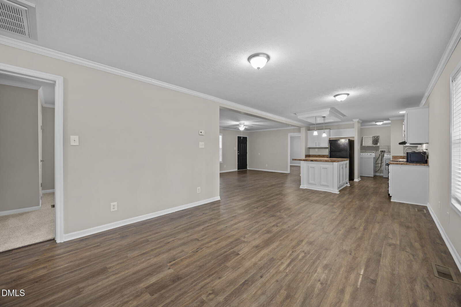 Open-concept living area with wood flooring, neutral walls, ceiling lights, and a kitchen with white cabinets and black appliances. A doorway leads to a carpeted room on the left.