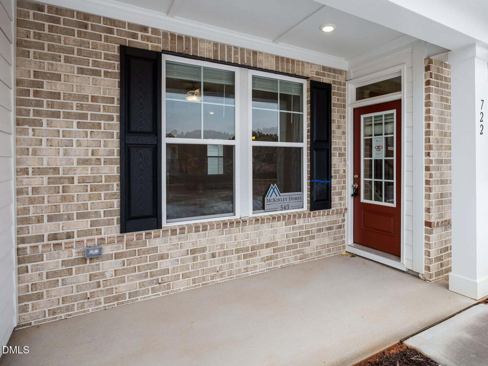 A brick house exterior with three large windows featuring black shutters and a red front door with glass panels. The porch has a covered ceiling and light fixture, and the house number 722 is visible on a white column.